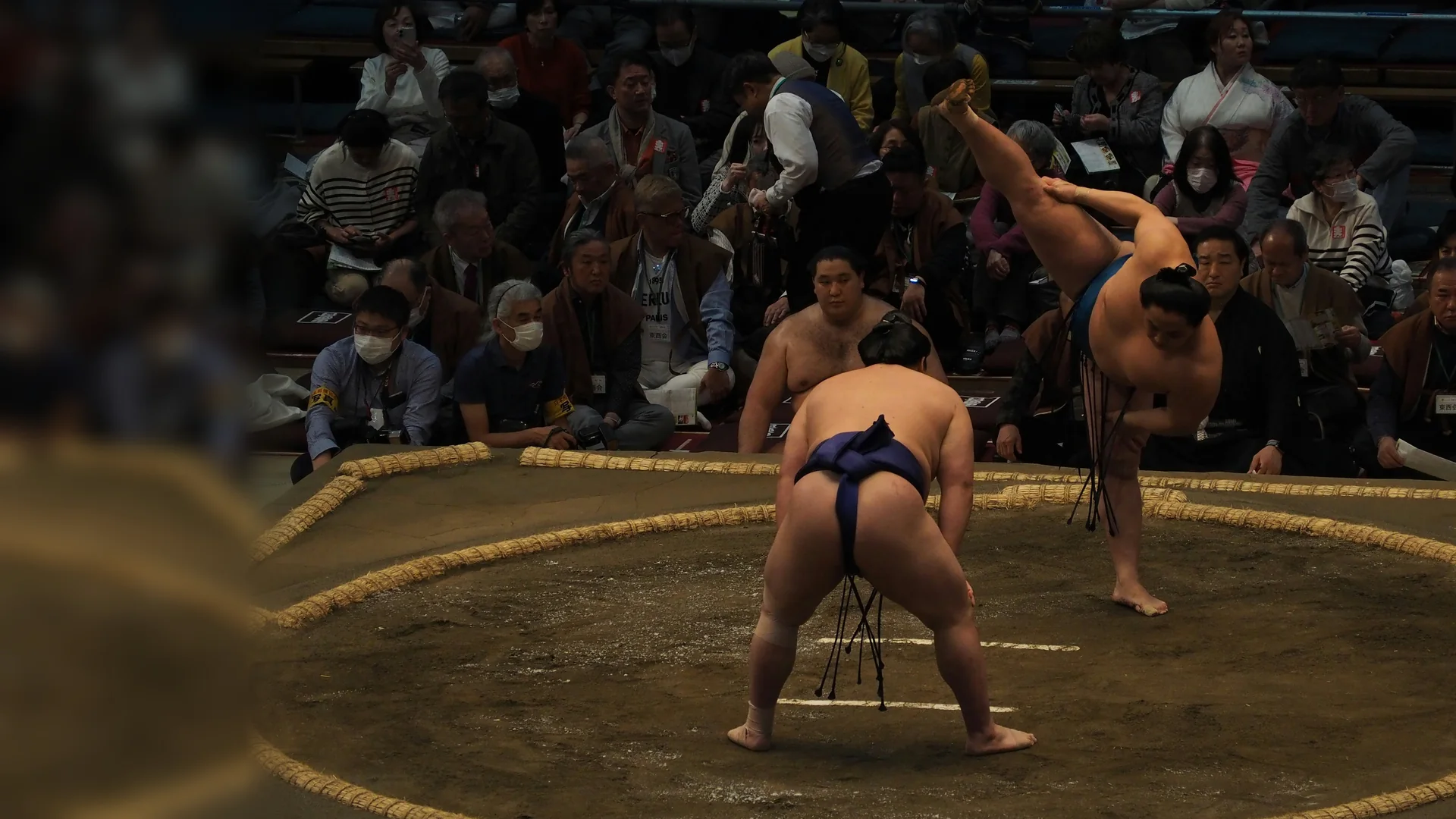 Two sumo wrestlers face each other inside a straw-bale ring, one with a leg raised in the ritual stamp, with spectators seated close around the clay
