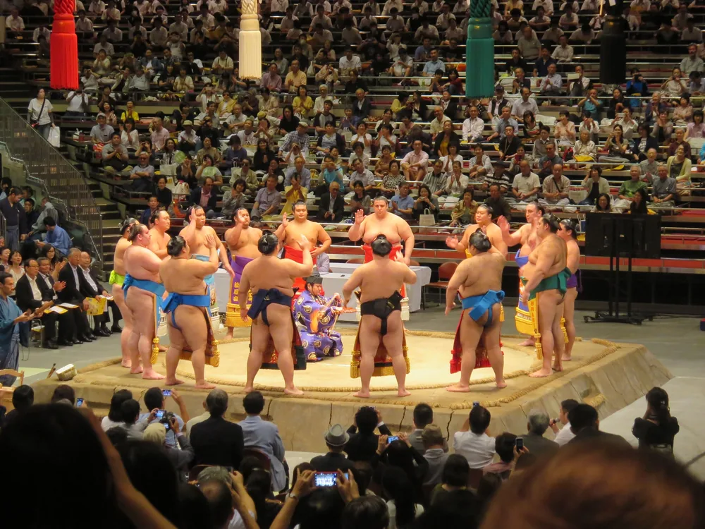 A circle of wrestlers in decorated ceremonial aprons stands on a raised clay ring around a referee at a professional tournament